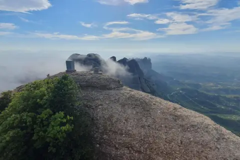 Cumbre rocosa con un monolito en Sant Jeroni, Montserrat. Nubes y montañas se extienden hasta el horizonte.