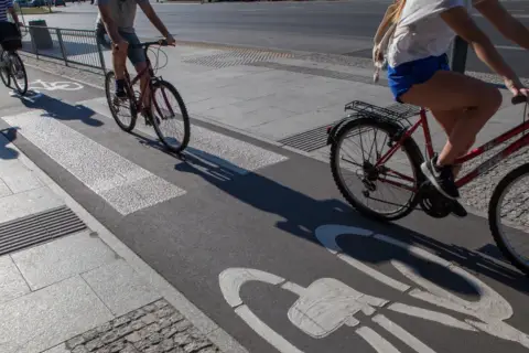 Ludzie na rowerach jadą ścieżką rowerową z oznaczeniem art 33 prd. Cienie rzucane przez rowerzystów wydłużają się na asfalcie.