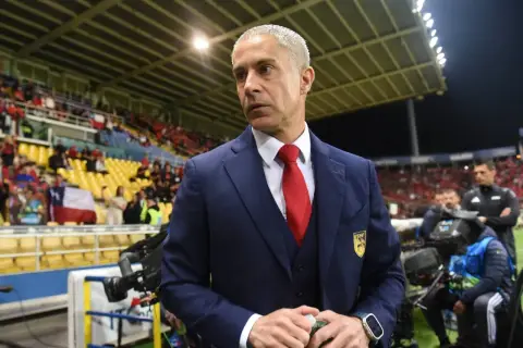 Sylvinho w granatowym garniturze i czerwonym krawacie, z emblematem na piersi, stoi na tle trybun stadionu.