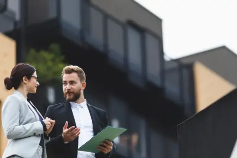 Dwóch agentów nieruchomości dyskutuje, jak wycenić mieszkanie, analizując dokumenty na tle nowoczesnego budynku.