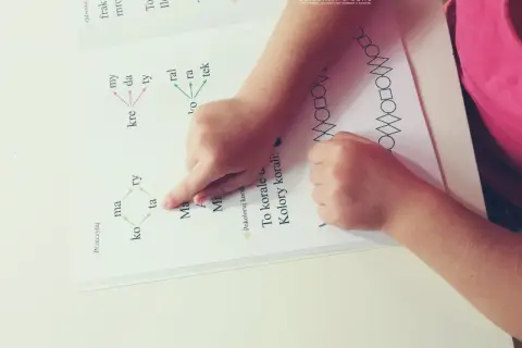 Dziecko ćwiczy czytanie, wskazując palcem na litery alfabetu i sylaby w książce. Obok widnieje wzór z figur geometrycznych.