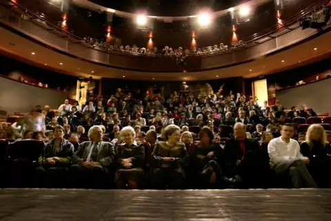 Widownia w teatrze, ludzie siedzą na miejscach, widać balkon z widzami.