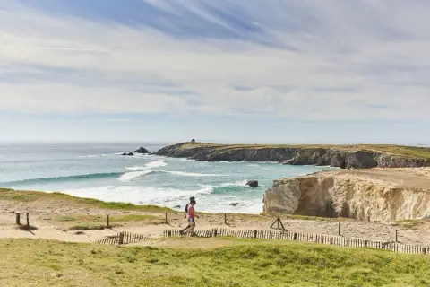 Deux personnes marchent sur un sentier côtier, découvrant les plus beaux endroits de Quiberon. Falaises dorées, mer turquoise et ciel bleu.