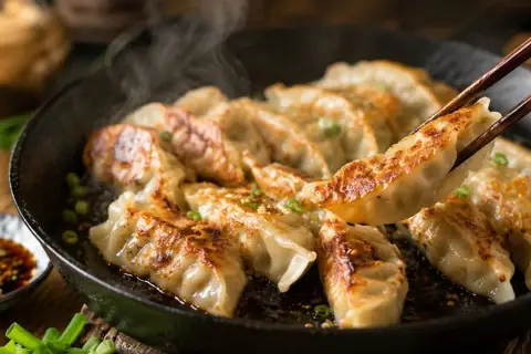 Gorące, rumiane pierożki gyoza parują na patelni. Jedna sztuka jest podnoszona pałeczkami, gotowa do zanurzenia w sosie.