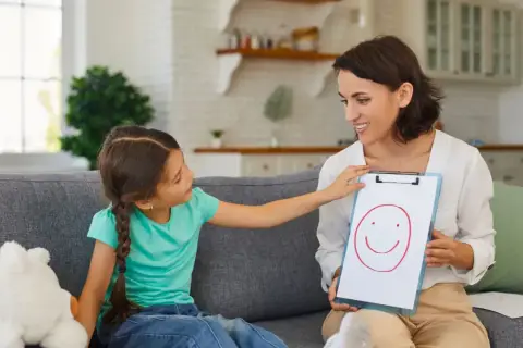 Logopedka pokazuje dziecku rysunek uśmiechniętej buźki na tablicy.