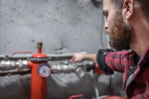 Uomo con barba controlla un manometro su un tubo rosso. Sento rumore nei tubi dell'acqua, forse un problema da risolvere.