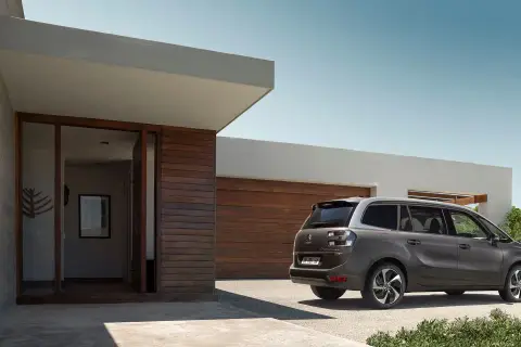 Szary minivan Citroën C4 Picasso zaparkowany przed nowoczesnym domem z drewnianą elewacją.