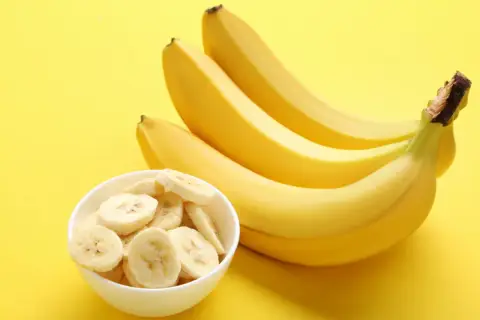 Pytanie: czy banany są kaloryczne? Na żółtym tle pęczek żółtych bananów i miseczka z plasterkami banana.