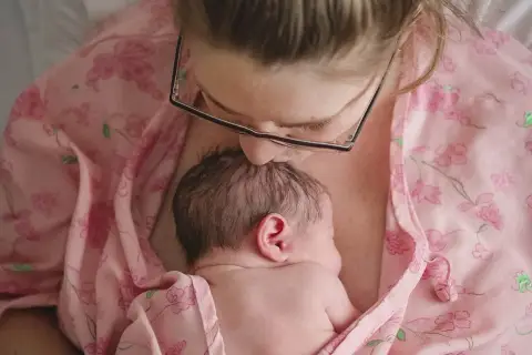 Mama całuje główkę noworodka, otulając go w różowy materiał. To piękny moment kangurowania, budowania więzi.