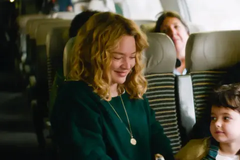 Una donna sorride mentre guarda un bambino su un treno. Le recensioni de "I figli degli altri" lodano questa scena.