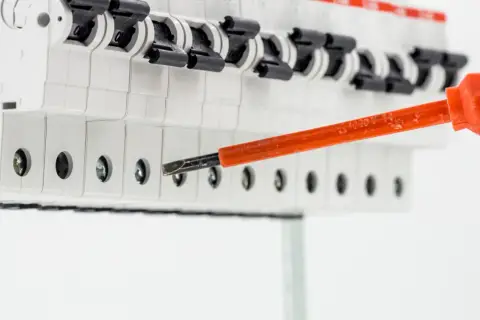 Elektryk dokręca śrubę w skrzynce z bezpiecznikami, przygotowując instalację. Widać przewód uziemienia i narzędzie rcd.