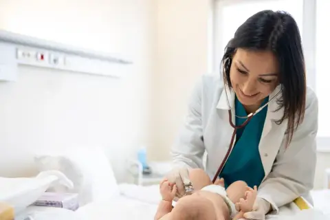 Lekarka w białym fartuchu osłuchuje stetoskopem noworodka leżącego na łóżku.