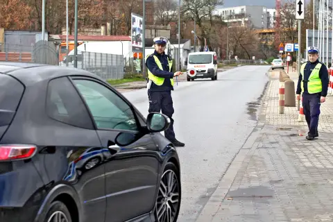 Policjant zatrzymuje czarne auto, być może za recydywę mandatu. Na drodze stoi biały van.