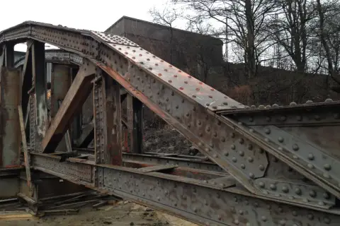Zardzewiały, nitowany most w awinionie, z widocznymi elementami konstrukcyjnymi i budynkiem w tle.