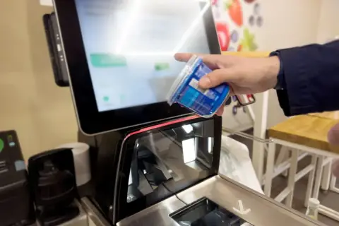 Ręka skanuje produkt przy kasie samoobsługowej. Ekran wyświetla informacje o zakupach i warunki płatności.