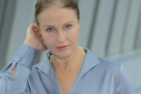 Portret kobiety w niebieskiej koszuli i perłach, opierającej dłoń o ucho.