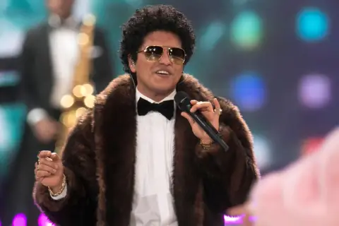 Bruno Mars w futrzanym płaszczu i okularach przeciwsłonecznych śpiewa na koncercie, trzymając mikrofon.