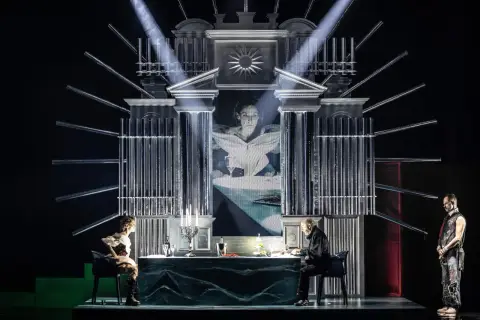 Scena teatralna z projekcją twarzy kobiety, dwójką siedzących przy stole postaci i stojącym mężczyzną. Atmosfera pełna jest teatralnych misteriów.