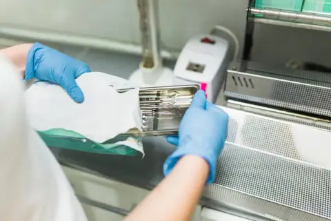 Ręce w niebieskich rękawiczkach wkładają tackę ze sterylnymi narzędziami do urządzenia, pokazując, jak działa autoklaw.