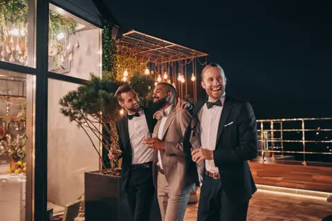 Trzech eleganckich mężczyzn w garniturach, z kieliszkami w dłoniach, śmieje się i rozmawia na tarasie. Jeden z nich, z brodą, jest wyraźnie w roli drużby.