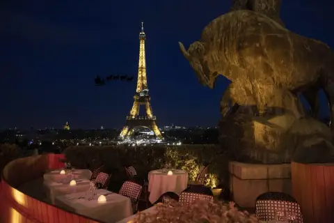 Vue nocturne de Paris avec la Tour Eiffel illuminée. Un bar ouvert le 25 décembre, avec des tables dressées et une statue de taureau. Le traîneau du Père Noël traverse le ciel.
