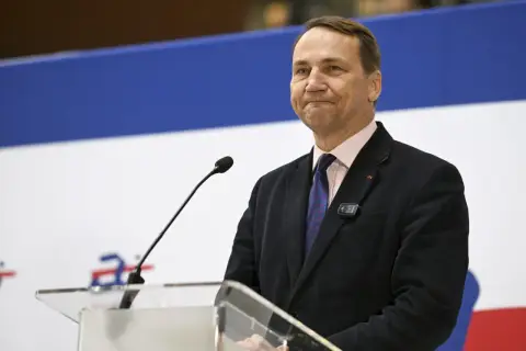 Mężczyzna w garniturze, podobny do Jana Sikorskiego, ojca Radosława Sikorskiego, przemawia przy mównicy.