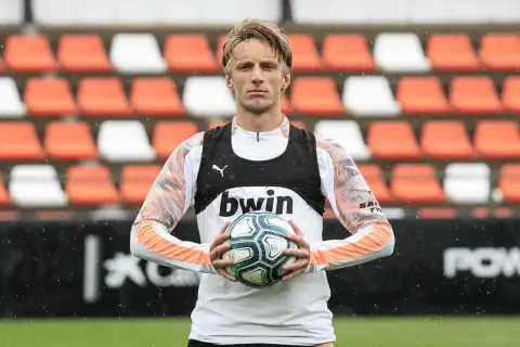 Piłkarz z blond włosami trzyma piłkę, ubrany w białą koszulkę z pomarańczowymi rękawami i czarną kamizelkę z logo bwin. W tle pomarańczowo-białe trybuny.