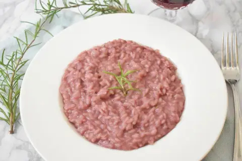 Aksamitne risotto z czerwonym winem, udekorowane gałązką rozmarynu, prezentuje się apetycznie na białym talerzu. Obok widoczny widelec i kieliszek wina.