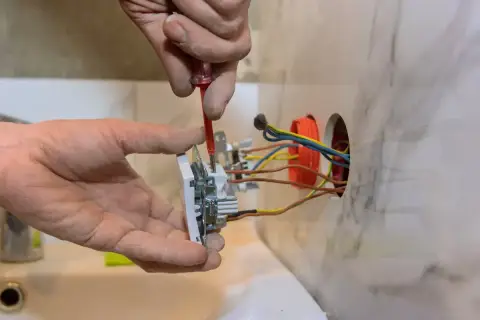 Ręce majsterkowicza montują gniazdko, przykręcając śrubki. W ścianie widać kolorowe przewody elektryczne, gotowe do podłączenia.