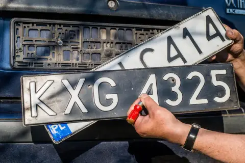 Wymiana tablic rejestracyjnych. Jedna tablica z numerem KXG 4325 jest przykręcana, druga z numerem C414 jest trzymana w ręku.