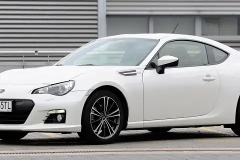 Białe Toyota BRZ z czarnymi felgami stoi przed budynkiem z poziomymi żaluzjami.