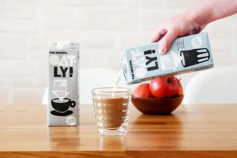 Ręka wylewa mleko dla baristów Oatly do szklanki. Obok stoi karton tego samego napoju.