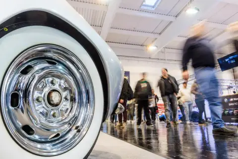 Detailaufnahme eines Oldtimer-Reifens mit Chromfelge auf einer motorsport expo. Besucher bewegen sich im Hintergrund.