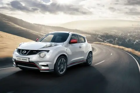 Biały Nissan Juke Nismo z czerwonymi lusterkami pędzi po krętej drodze z widokiem na miasto w oddali.