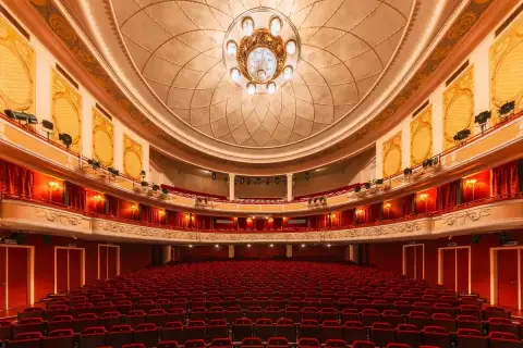 Wnętrze teatru z czerwonymi fotelami i bogato zdobionym sufitem. Czekamy na wspaniały występ.