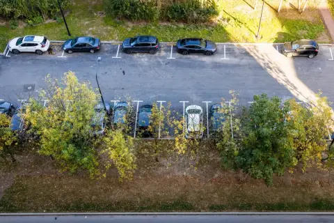 Widok z góry na parkowanie samochodów dostawczych na osiedlu. Kilka aut stoi na wyznaczonych miejscach, otoczonych zielenią i drzewami.