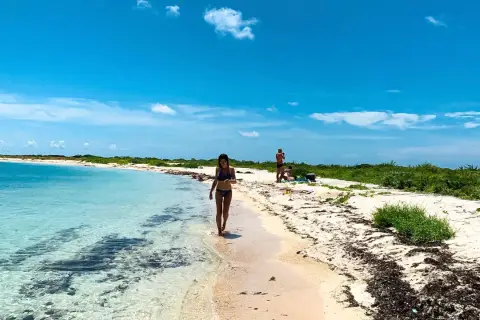 Kobieta spaceruje brzegiem turkusowej wody na plaży w Zatoce Meksykańskiej. W tle widać innych plażowiczów i bujną zieleń.