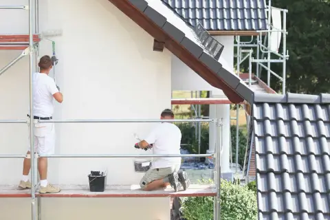 Dwóch malarzy na rusztowaniu odnawia elewację domu. Używają wałków i wiadra z farbą malarską rozpuszczalną w wodzie.