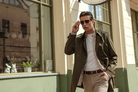 Mężczyzna w eleganckim, khaki płaszczu i beżowych spodniach poprawia okulary, idąc ulicą.