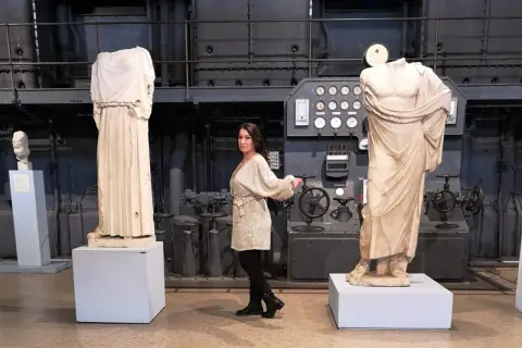 Donna in abito scintillante tra statue classiche e macchinari industriali, forse nel vecchio gasometro.