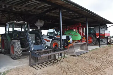 Ciągniki i maszyny rolnicze pod zadaszeniem. Widać zielony Fendt z ładowaczem, biały Bobcat, zielony opryskiwacz i ciągnik z rozsiewaczem. To wszystko w budynku gospodarczym bez zgłoszenia.