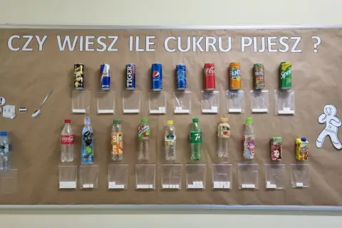 Tablica edukacyjna pokazuje, ile cukru jest w popularnych napojach. Obok każdej butelki i puszki znajduje się woreczek z odpowiadającą ilością kostek cukru.