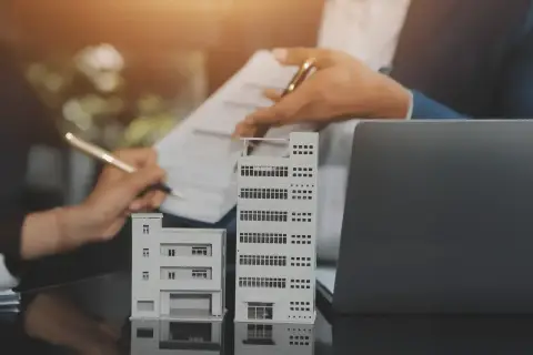Dwie makiety budynków, jedna niższa, druga wyższa, obok laptopa. Dłonie podpisują dokumenty, sugerując transakcję lub planowanie inwestycji, być może związanej z pcc 3.