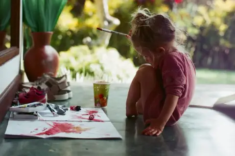 Kleines Mädchen malt mit Pinsel und Farben, um orange zu mischen. Bunte Tupfer auf Papier, ein Becher und Schuhe daneben.