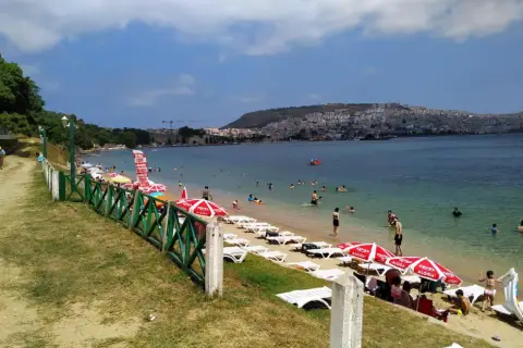 Plaża z leżakami, parasolami i ludźmi kąpiącymi się w morzu, w tle miasto na wzgórzu.