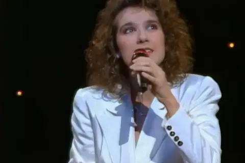 Celine Dion w białej marynarce śpiewa do mikrofonu, przypominając jej występ na Eurowizji.