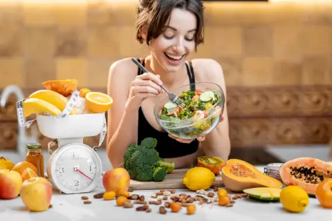 Mujer sonriente come ensalada. Alrededor, frutas, verduras y una báscula, sugiriendo una tabla de dieta para engordar.