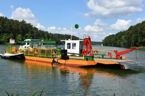 Prom na Zalewie Prężyce z białym vanem i pomarańczowym pokładem, otoczony zielonymi lasami.