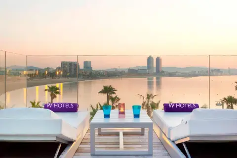 Dos tumbonas blancas con toallas moradas de W Hotels, una mesa con vasos y velas, y vistas al mar y la ciudad de Barcelona al atardecer.