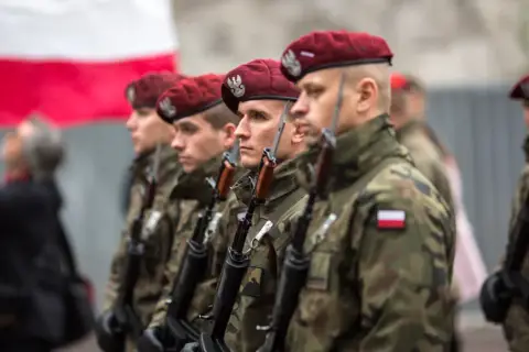 Żołnierze w bordowych beretach i mundurach polowych, z karabinami, reprezentują różne kategorie wojskowe. W tle biało-czerwona flaga.